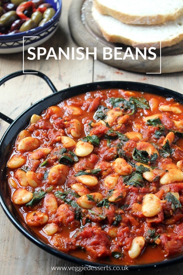 Spanish Beans with tomatoes in a black dish, which can be served as a main meal or tapas. Photographed with bread and olives. Vegan and gluten free recipe.