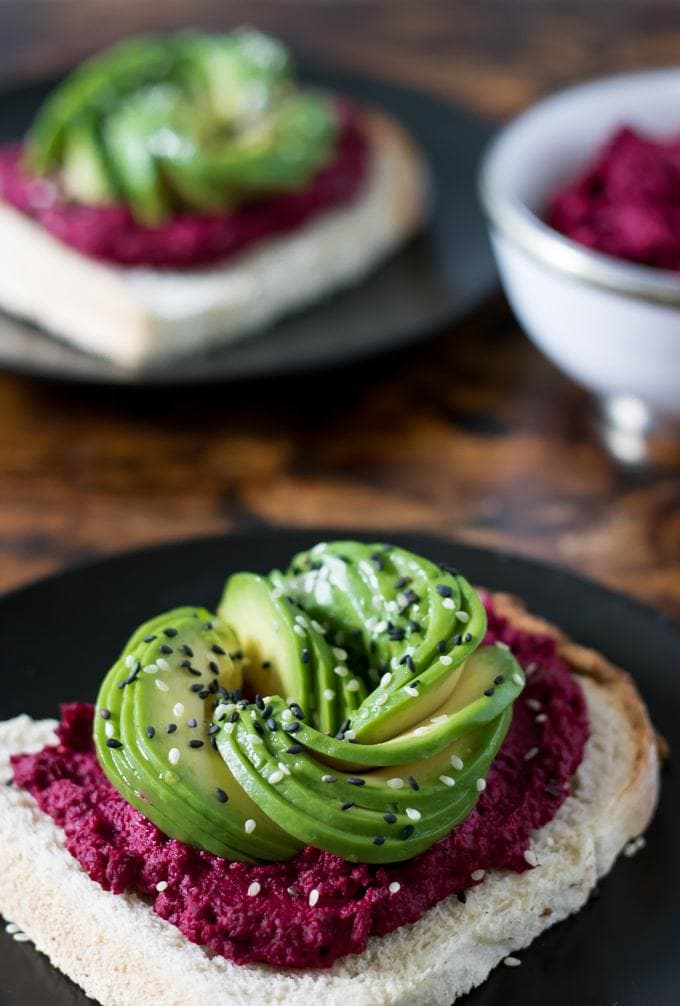 Close up of toast spread with beet hummus and topped with avocado sliced into a rose shape, then sprinkled with black and white sesame seeds.