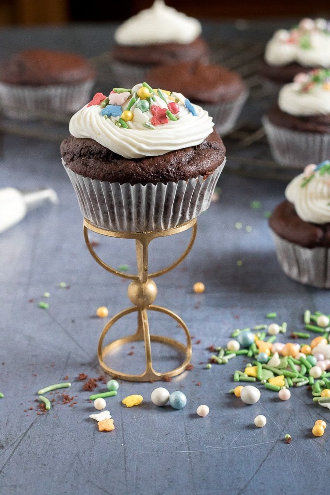 A chocolate vegan cupcake on a golden cupcake stand with vanilla icing and sprinkles. Get the easy vegan chocolate cupcakes recipe now!