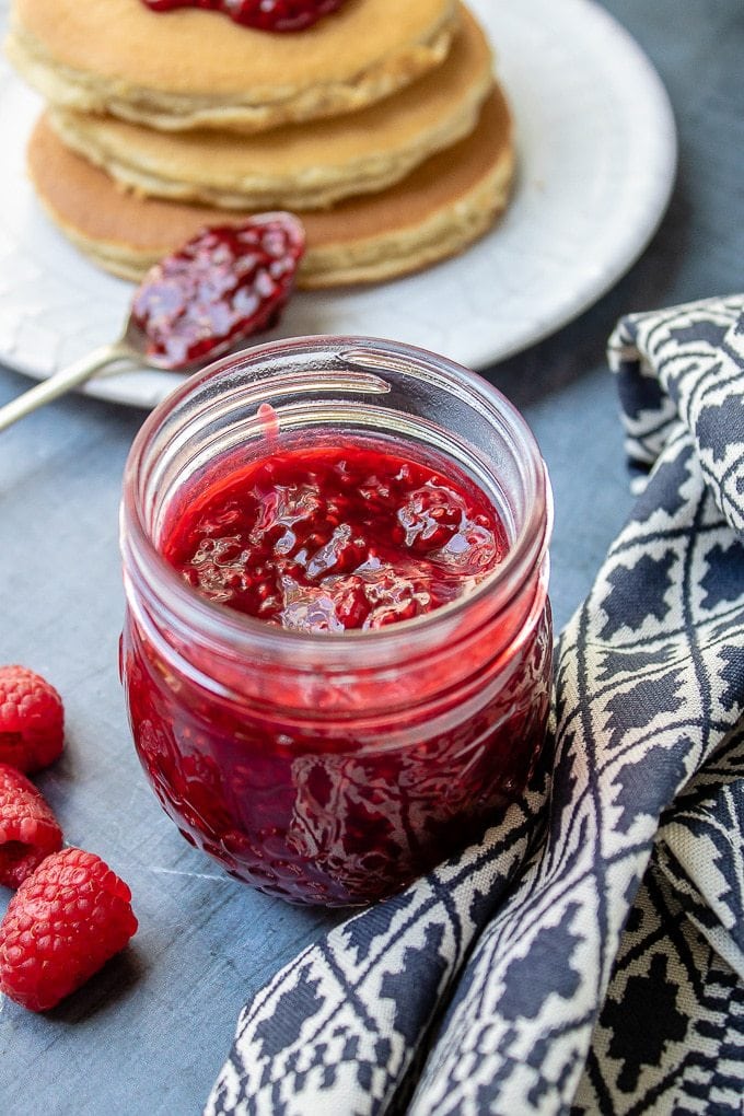 Close up of a jar of thick raspberry compote next to a stack of pancakes to spoon it onto.