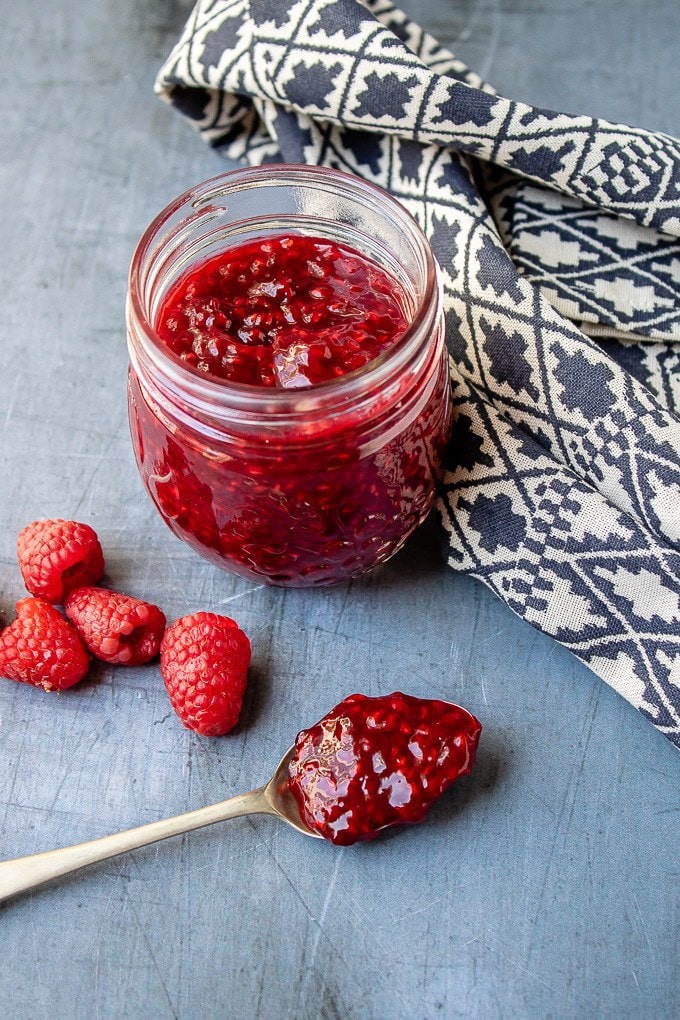 A jar of quick, easy and tasty raspberry compote, with a spoonful of it next to a blue and white tea towel.