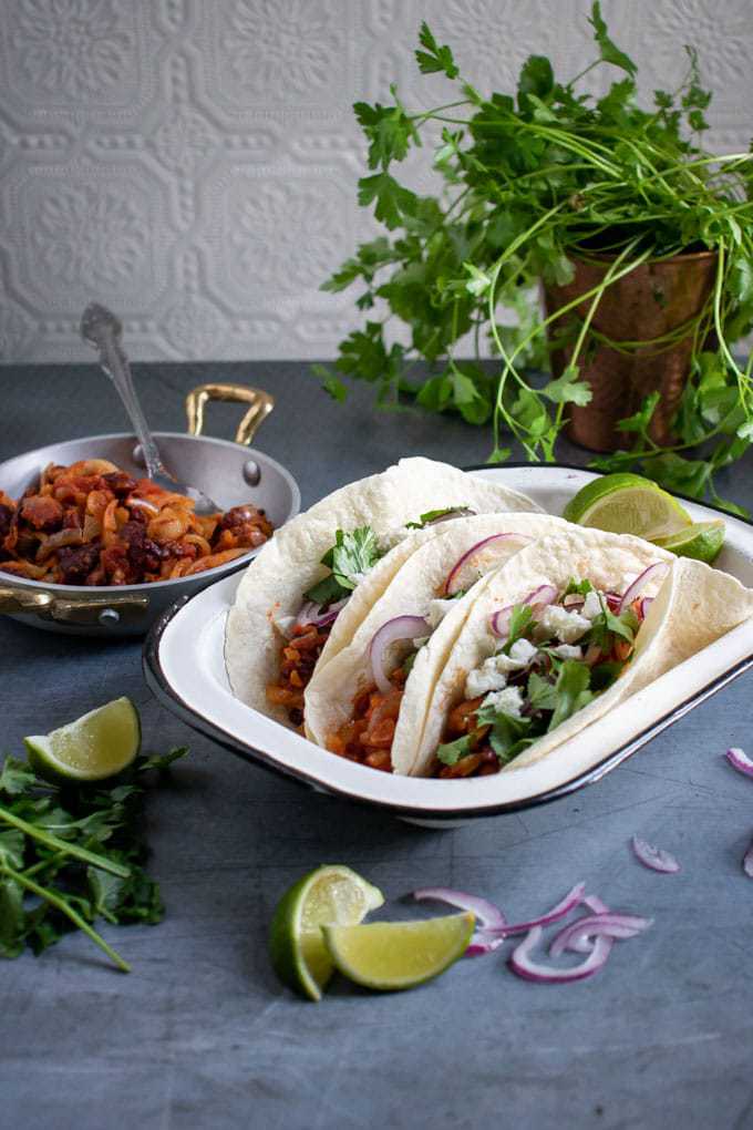 Enamel dish with three vegetarian tacos - made in a Tefal Actifry air fryer. In front of fresh coriander, limes and red onion.