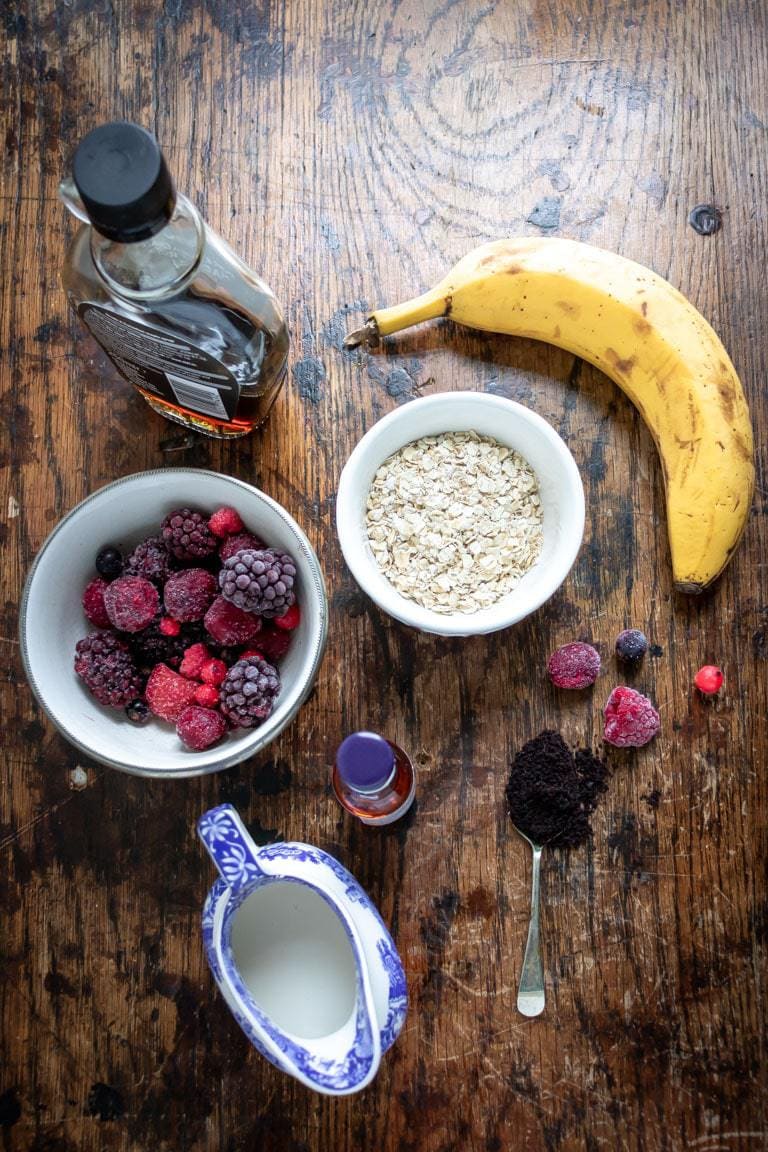 Ingredients for acai smoothie bowl recipe on a wooden table - banana, oats, acai powder, plant milk, vanilla, maple syrup, frozen berries.