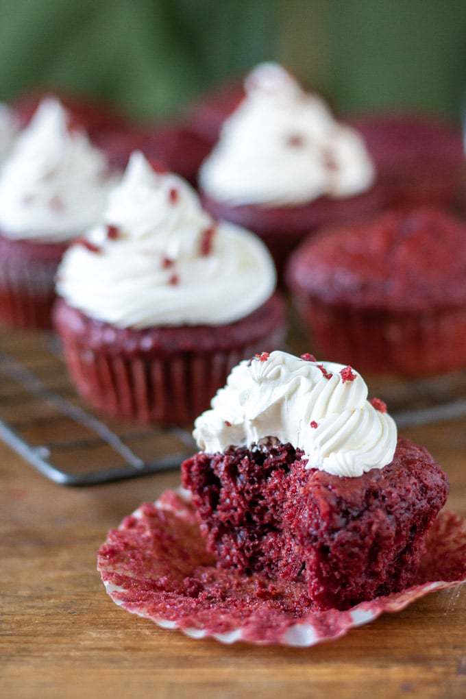 Close up of a vegan red velvet cupcake with vegan cream cheese frosting with a bite taken out in front of a rack of cupcakes.