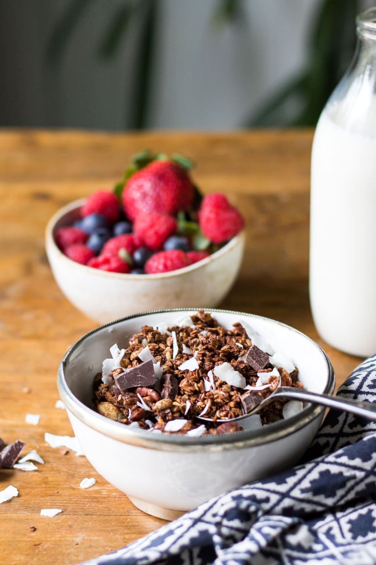 A bowl of homemade granola. The vegan granola with chocolate and coconut is shown in front of a bowl of berries.