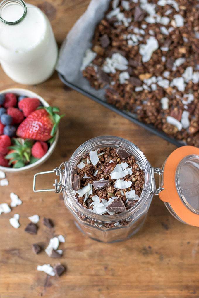 A glass jar full of fresh chocolate granola with coconut. Next to a bowl of berries.