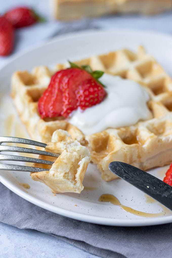 Close up of a fork with a piece of fluffy vegan waffle, topped with dairy free yogurt and strawberries.