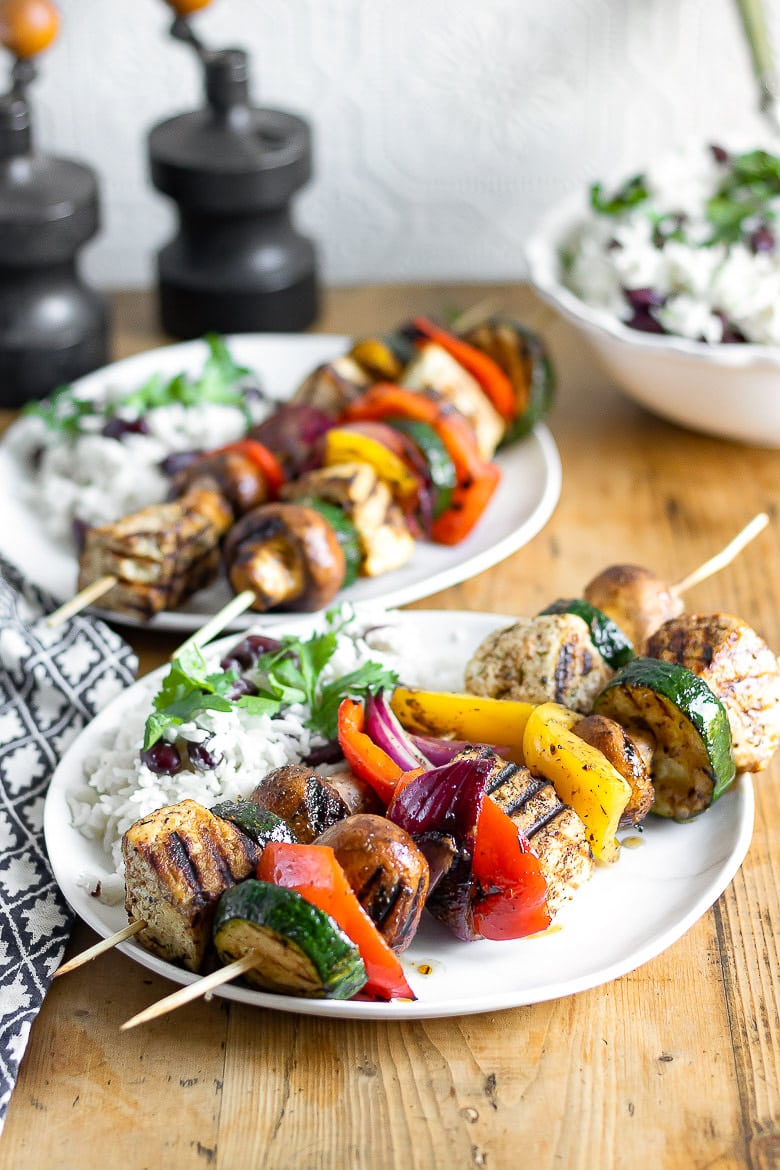 Close up of grilled jerk tofu on skewers with vegetables, shown on a plate with Jamaican rice and peas