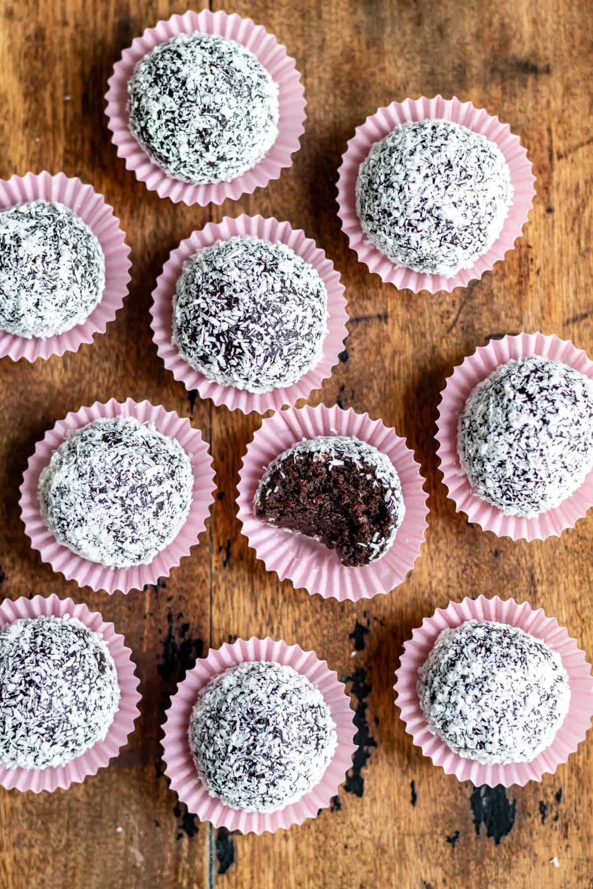 Rows of romkugler Danish rum balls in pink paper wrappers on a wooden table, one has a bite out.