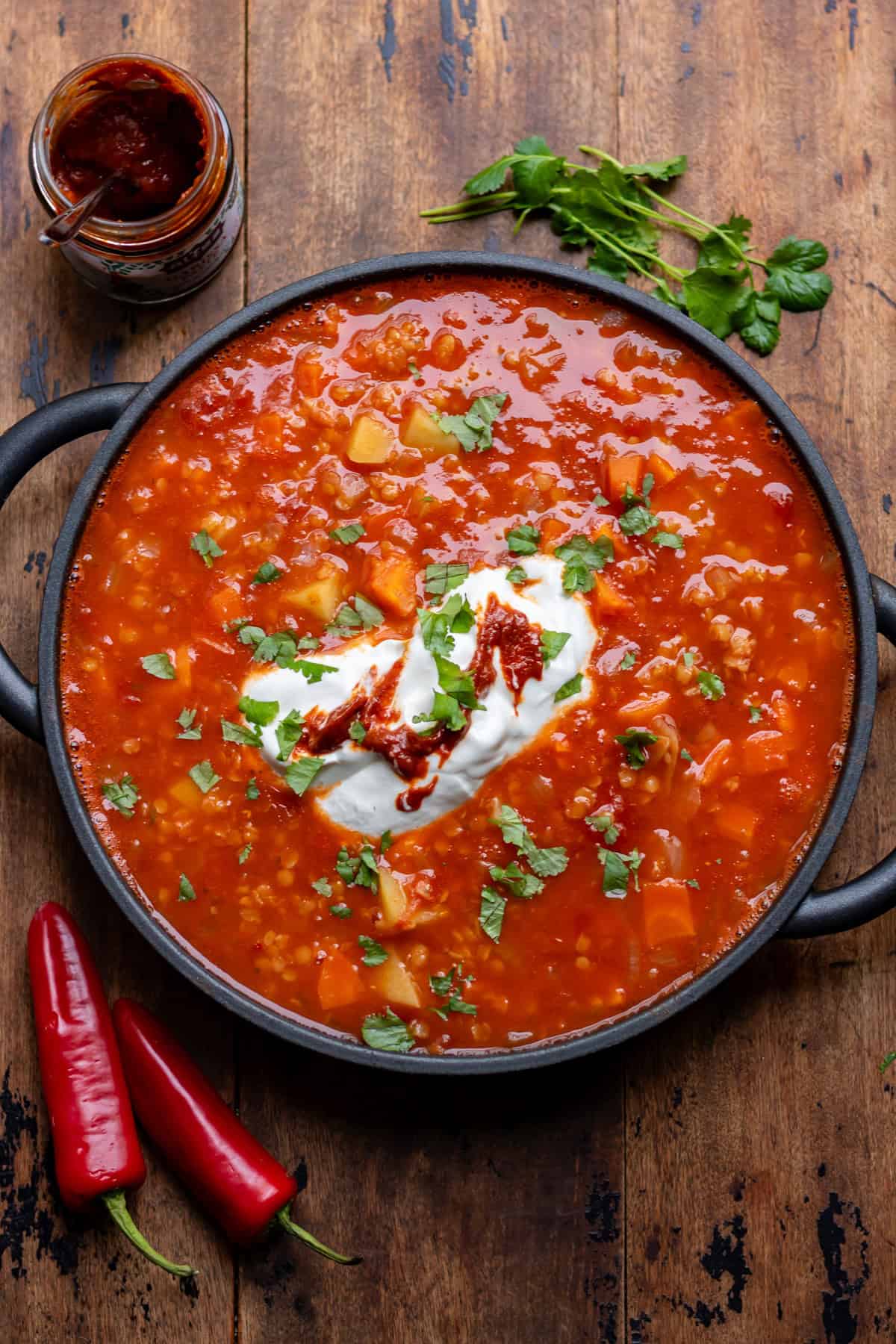 A wooden table with a serving dish of tomato lentil soup, topped with yogurt and harissa paste.