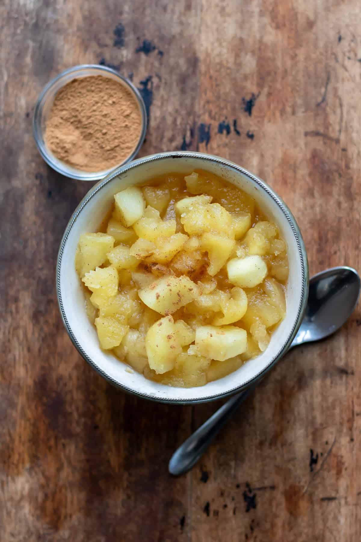 A wooden table with a dish of apple compote and a small dish of cinnamon.