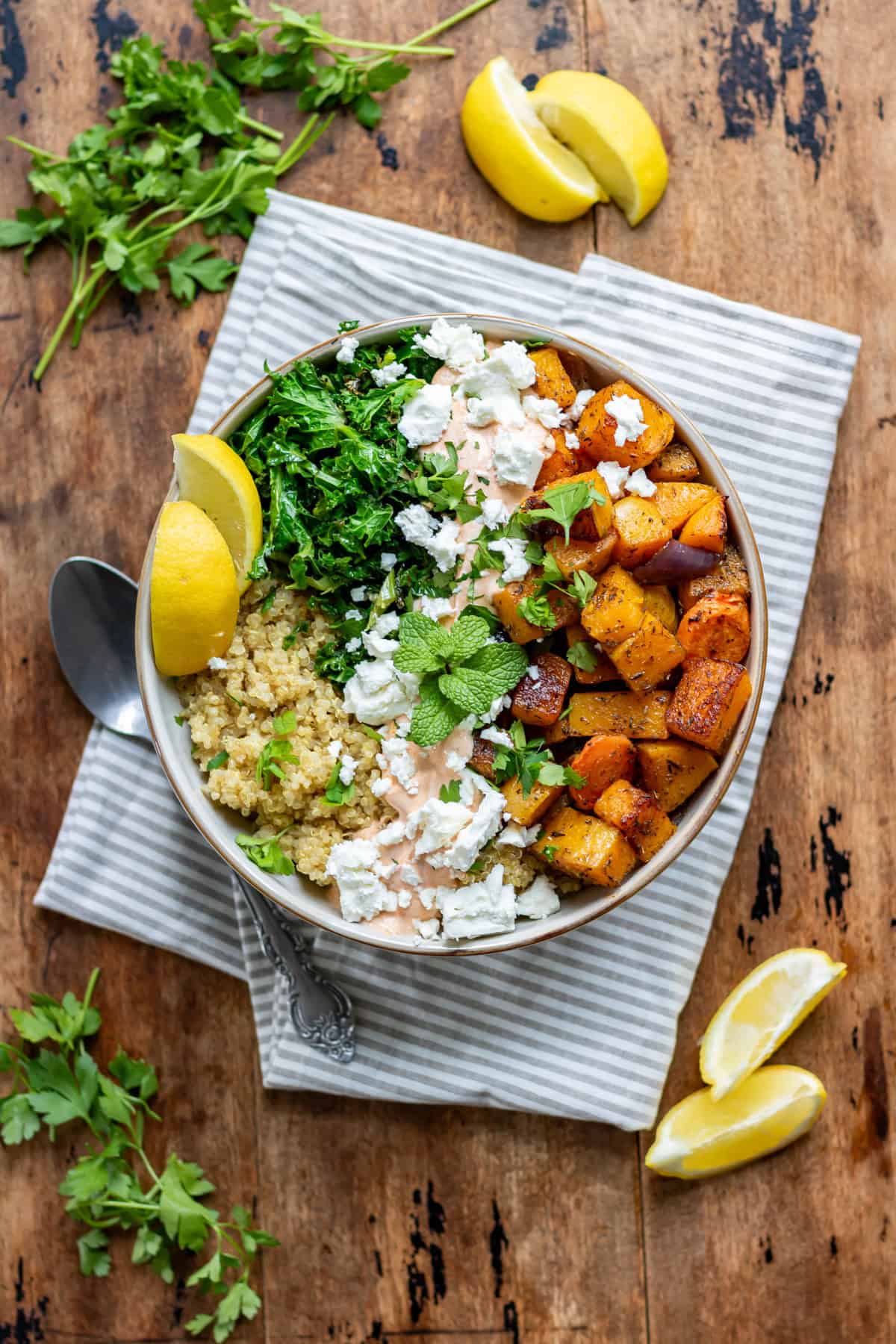 A wooden table with a bowl of roasted vegetable quinoa salad, with parsley and sliced lemons scattered around.