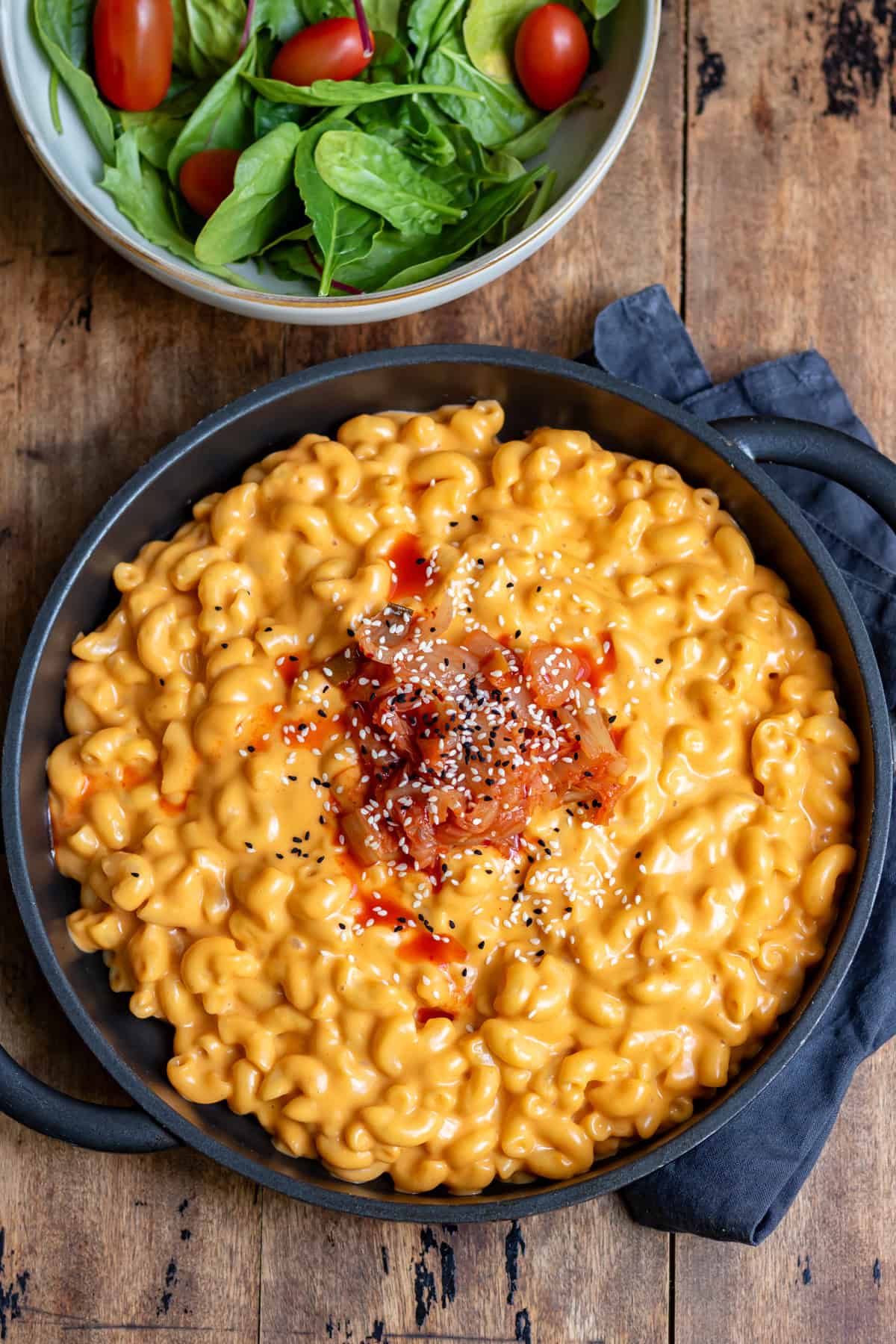 A wooden table with a serving dish of Korean gochujang mac n cheese, next to a dish of salad.
