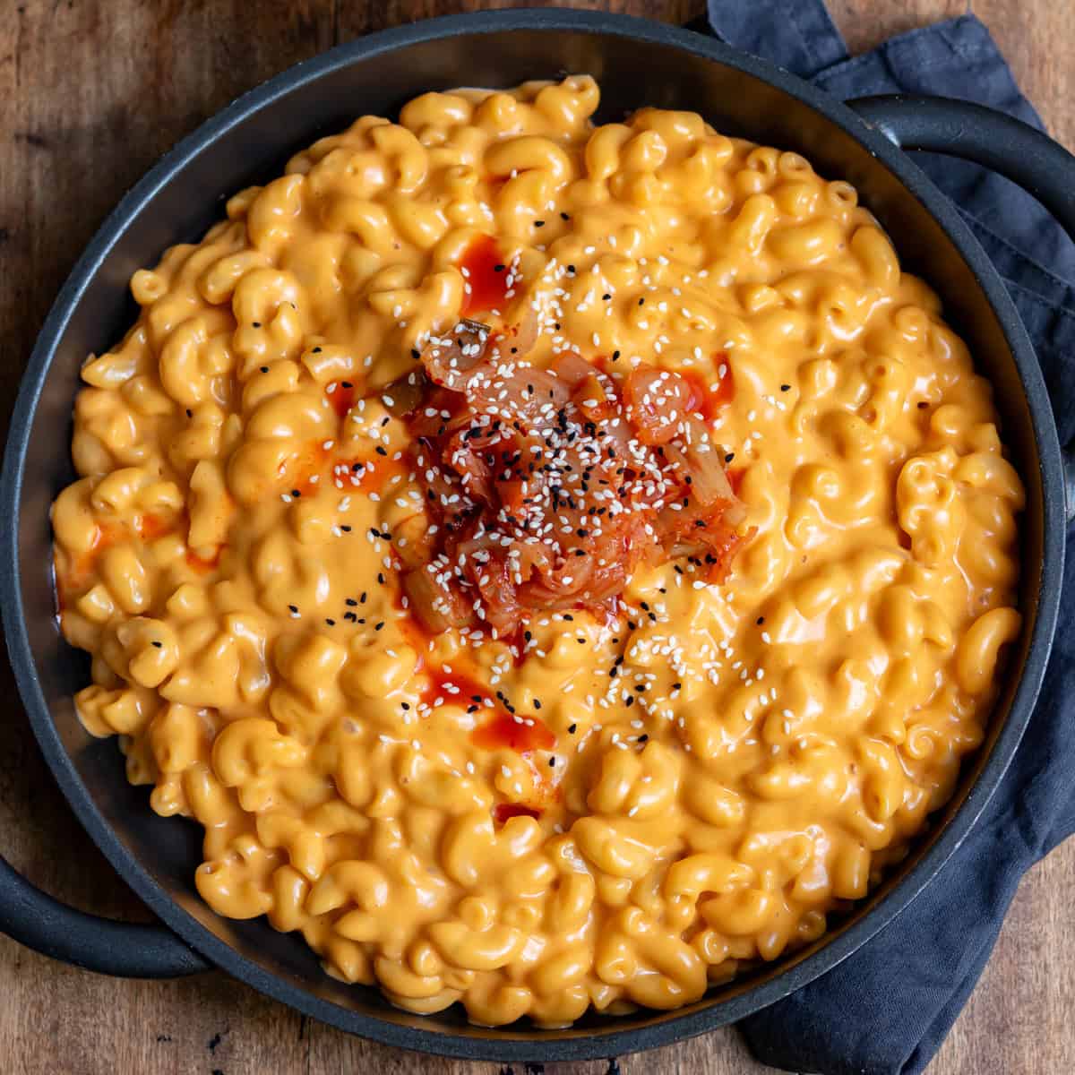 A wooden table with a serving dish of Korean gochujang mac n cheese, topped with kimchi.