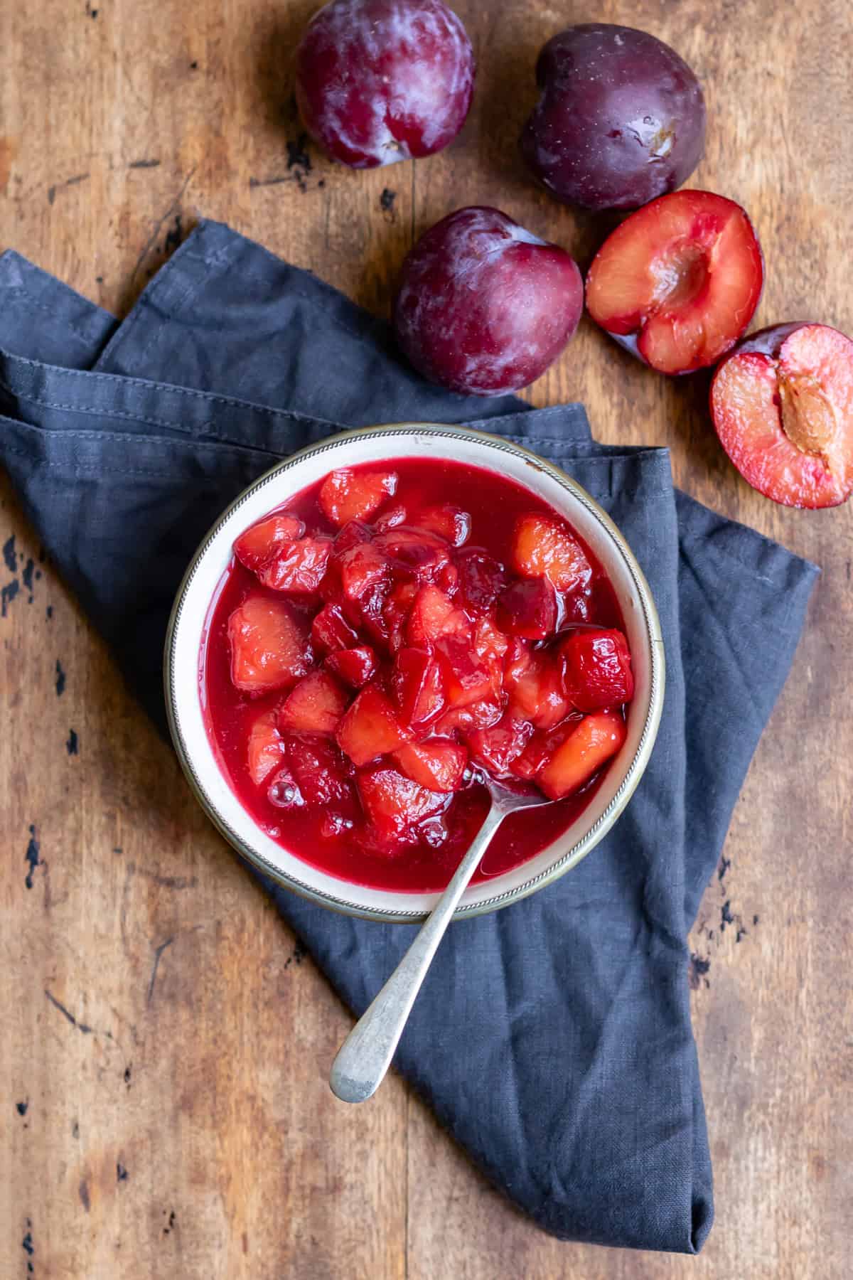 A wooden table with a bowl of plum compote, plus plums next to it.
