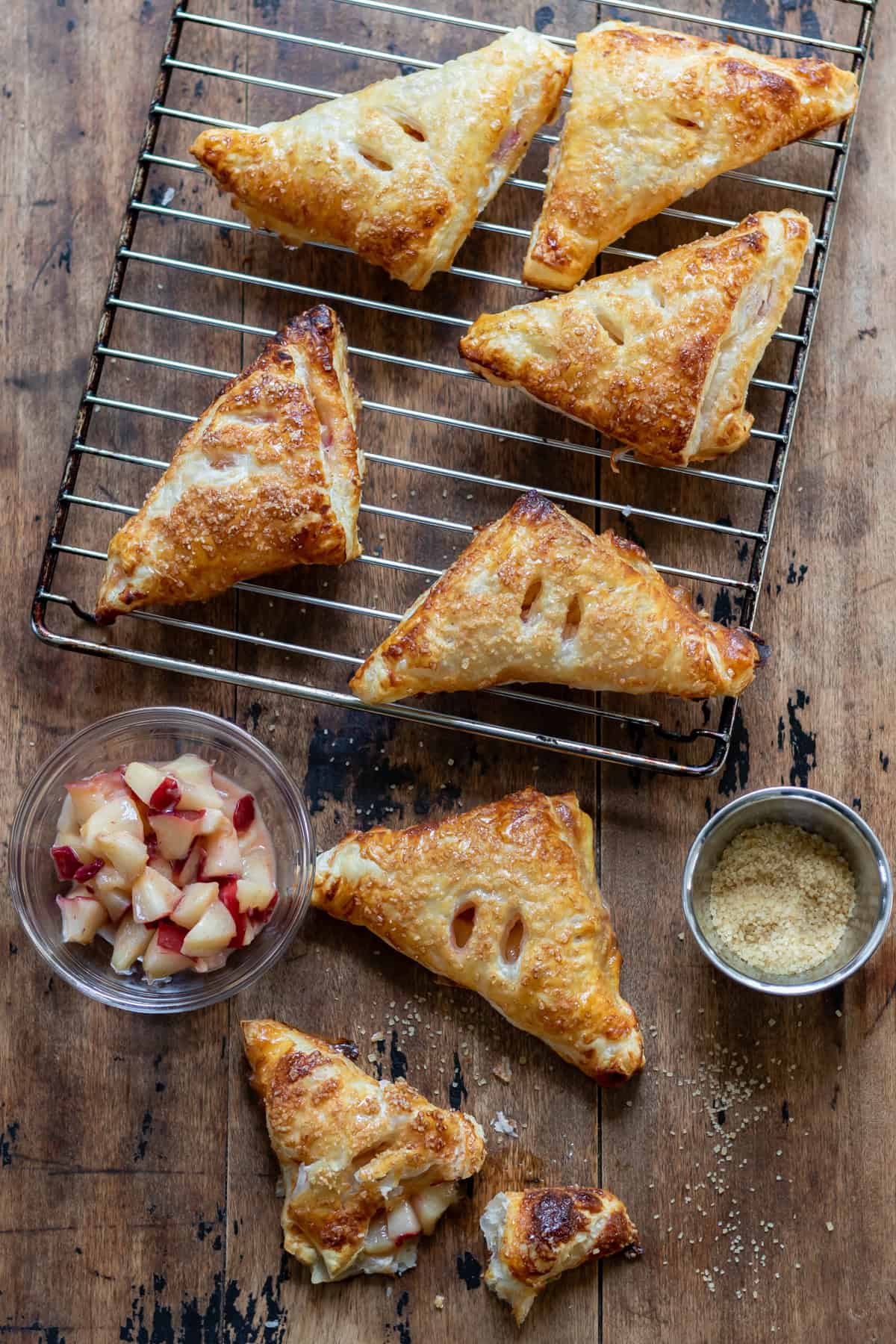 A wooden table with a rack of cooling nectarine turnovers, a few turnovers on the table, a dish of nectarines and a dish of coarse sugar.