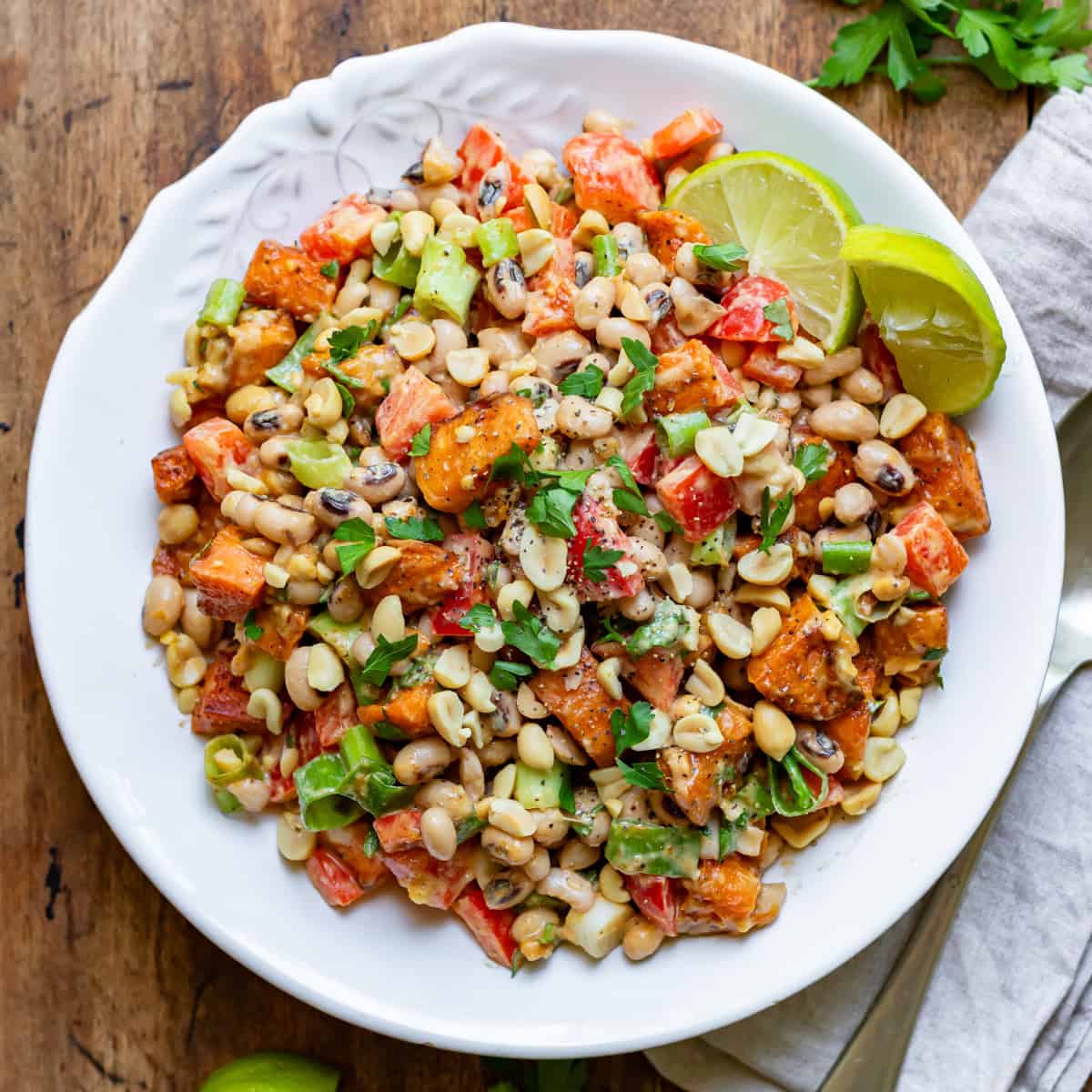 Looking down at a wooden table with a serving dish of sweet potato black eye peas salad with peanut dressing.